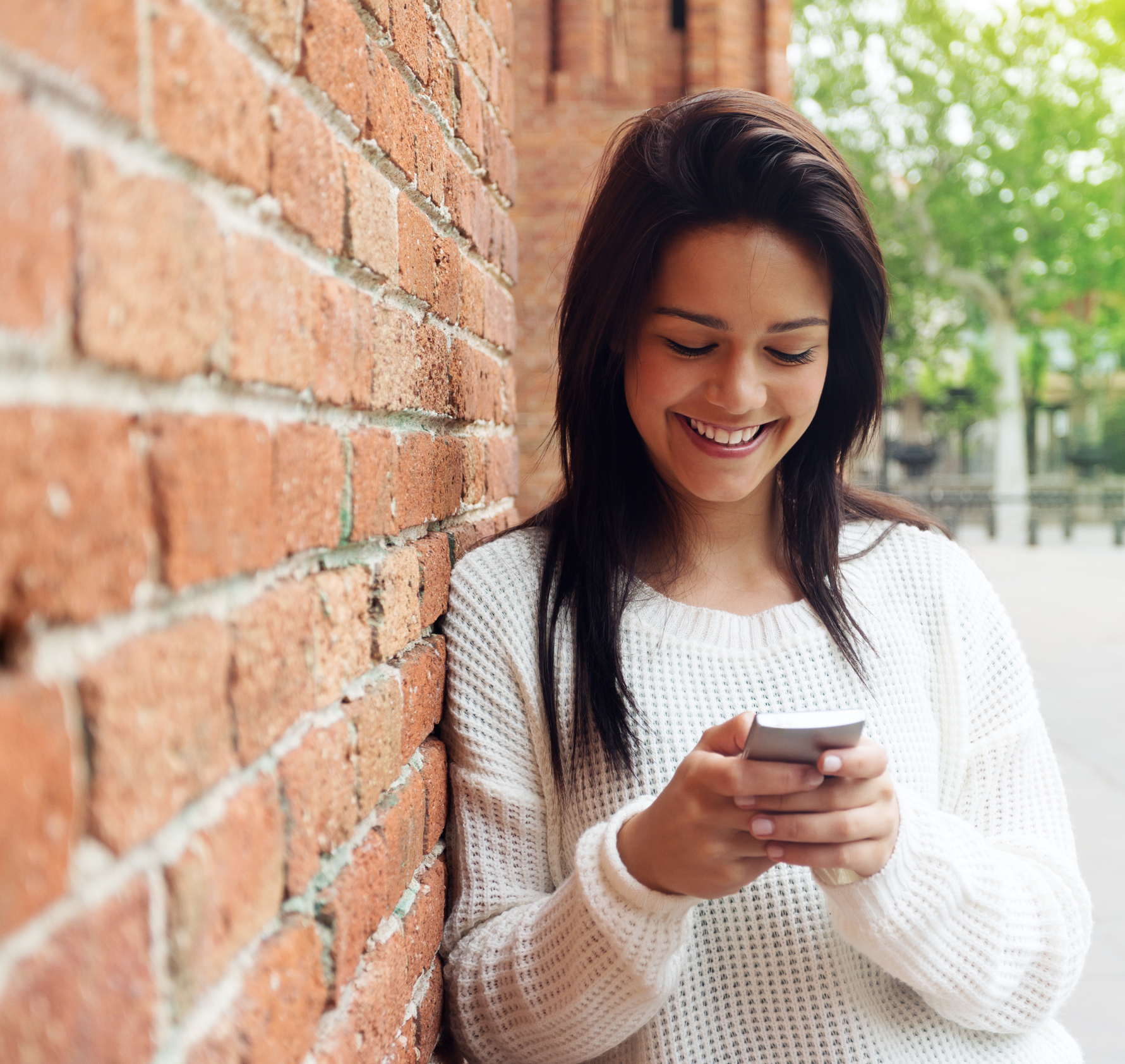 Beautiful woman using the smartphone sending a text message with happy expression in front of a wall. Natural light. Square photography from a DSLR camera. Sharp focus.