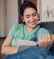 A young woman in overalls reading a book on a sofa.